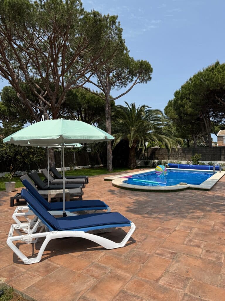 Terrasse mit mehreren Liegestühlen unter einem Sonnenschirm neben einem rechteckigen Swimmingpool, umgeben von hohen Bäumen und einem klaren Himmel im Hintergrund.