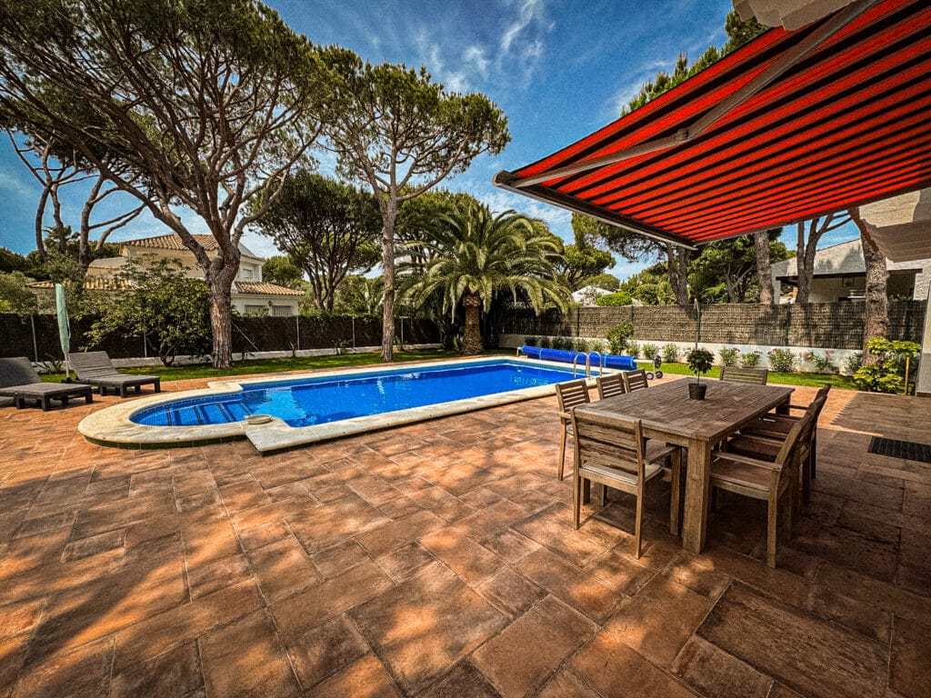 Garten mit Swimmingpool, hölzernem Esstisch, Stühlen und einer Markise in Rot über einem Terrassenbereich; umgeben von hohen Bäumen und Sträuchern unter einem blauen Himmel.